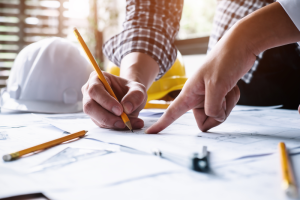 Close-up of two hands at a desk reviewing architectural plans. One hand holds a pencil, the other points. Hard hats and tools suggest a construction setting.