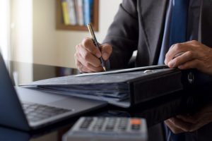 A person in a suit writes in a binder on a reflective desk, near a laptop and calculator, conveying focus and professionalism in an office setting.