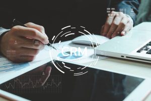 Two hands work with documents and a laptop. The word "CRM" and digital icons float above, suggesting customer relationship management in a business setting.