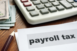 Calculator and cash on a wooden table beside a document labeled "payroll tax," conveying financial management and taxation themes.