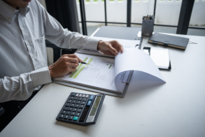 A person examines financial documents in a binder at a desk. A calculator, notebook, and stationery are nearby, conveying a focused, professional tone.